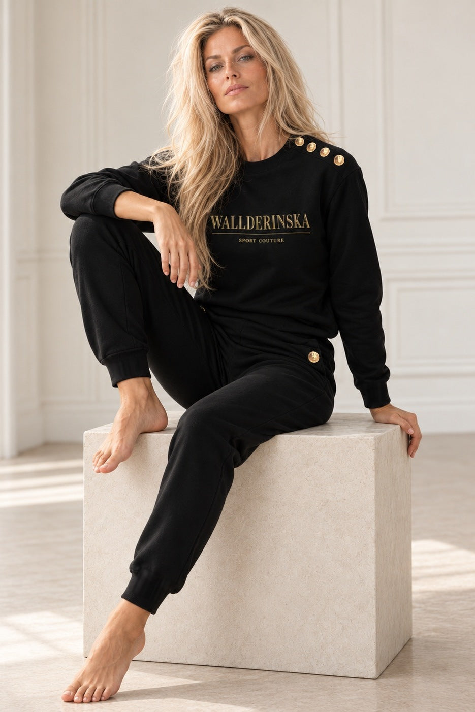 Edge Gold Button Sweater och Pants i svart, modellen sitter på en ljus stenkub i vit studio, med guldknappar på axeln och vid fickan.