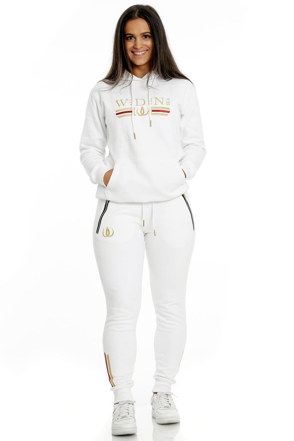 Stående i hvide joggers med guldlogo og hvid hættetrøje med guldbroderi, til hvide sneakers.
