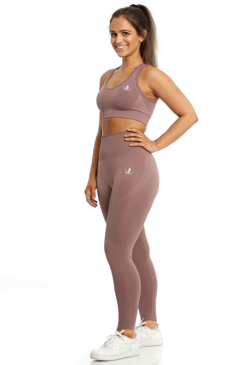 Sports-bh og tights i samme rosabrune tone. Modellen står med hånden i siden, i hvide sko.