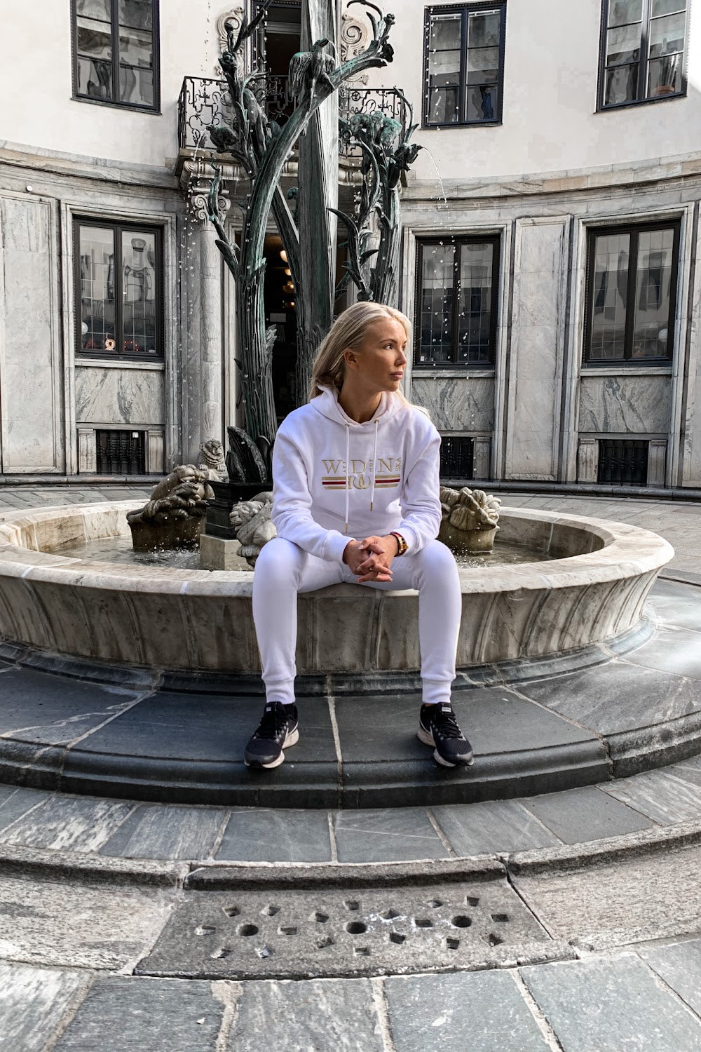 På springvandskanten sidder modellen i den hvide hættetrøje, med en statue i baggrunden.