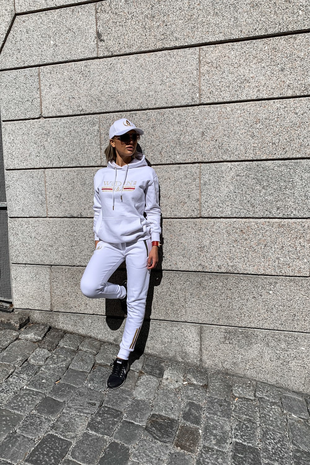 Op ad stenmuren læner modellen sig med den ene fod trukket op, i hvid hættetrøje og hvide joggers.