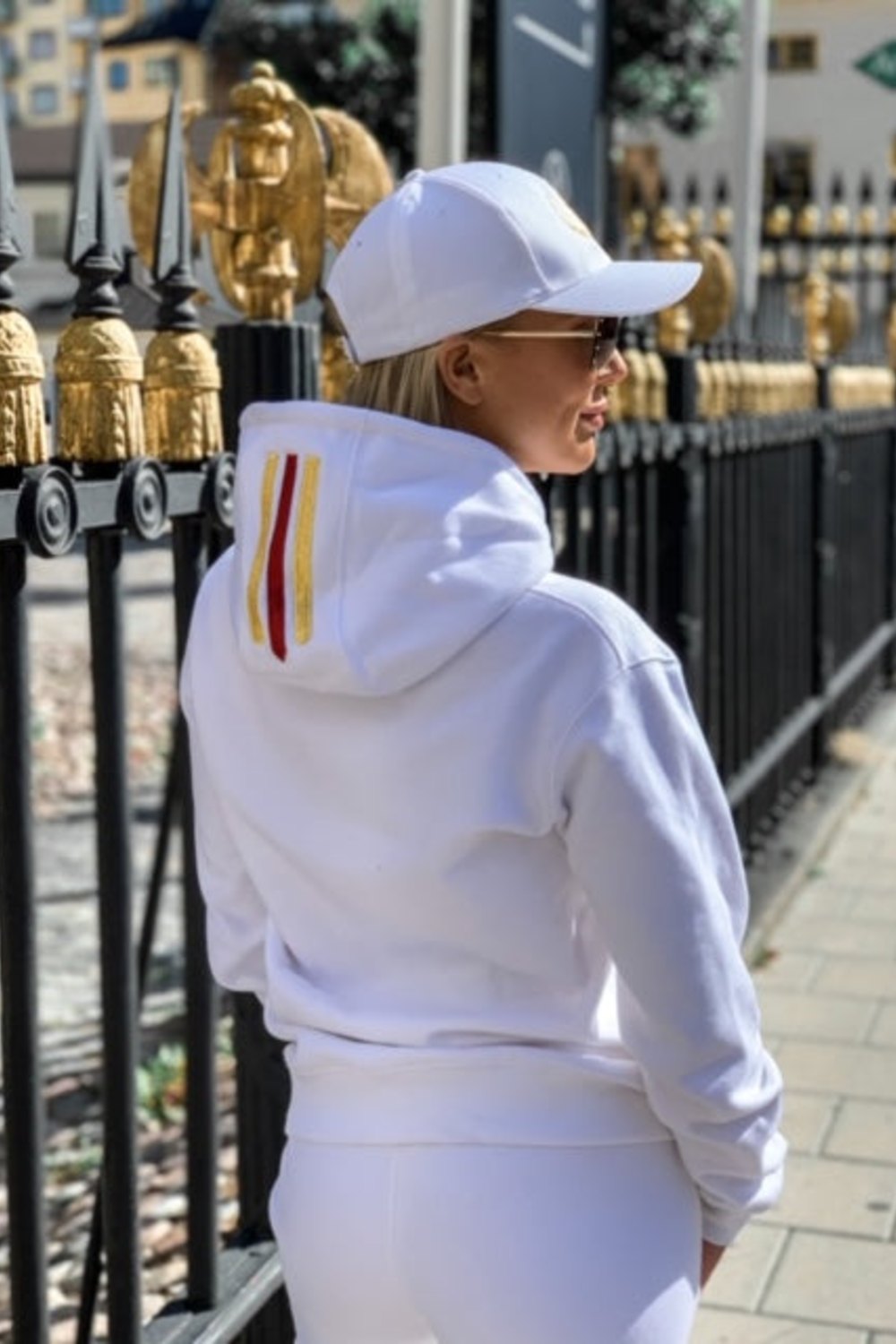 Guld og rød stribe på hætten ses, når modellen står vendt mod et sort hegn.