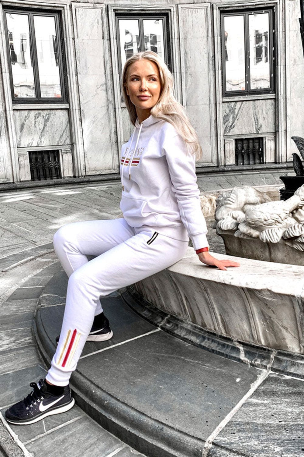 Modellen sidder ved et springvand i byen i den hvide hættetrøje og hvide joggers med guldstribe.