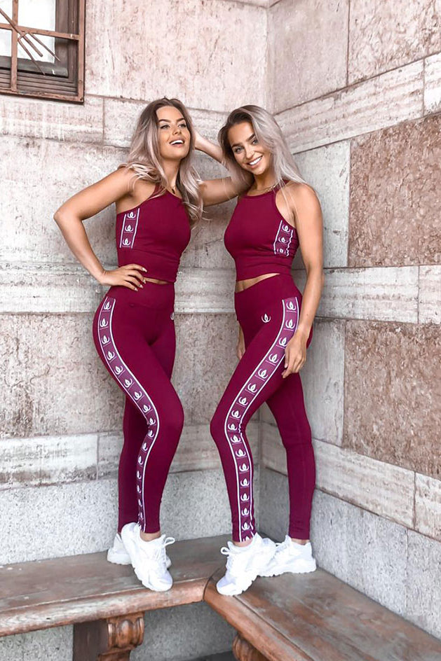 To modeller lænet op ad en stenmur i vinrøde croptops og tights med hvide logobånd.
