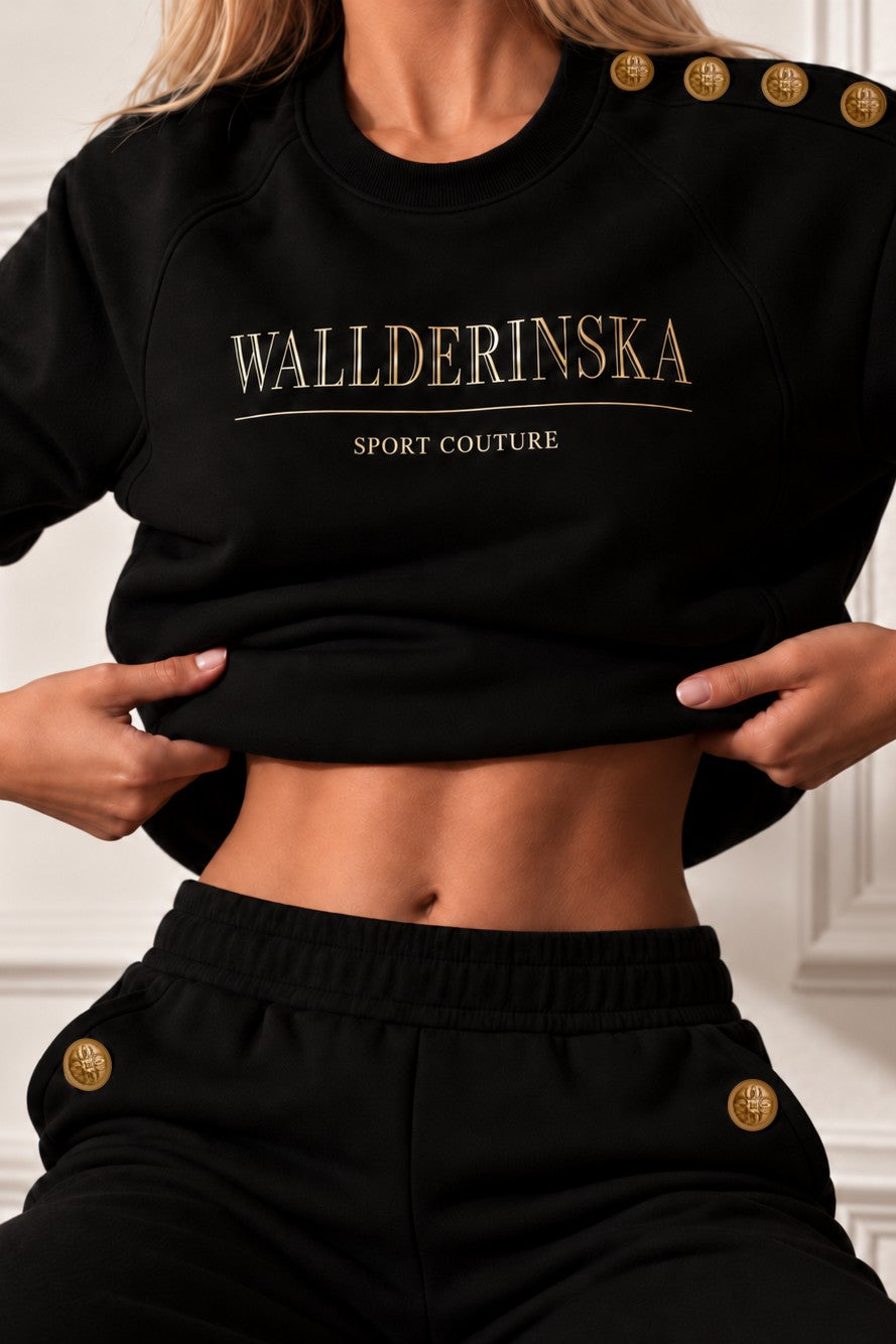 Edge Gold Button Bukser i sort med guldknapper ved lommerne, og sweateren løftet over taljen.
